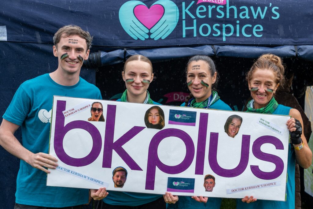 Three women and one man in front of a Dr Kershaw's Hospice banner holding a sign saying BK Plus