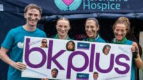 Three women and one man in front of a Dr Kershaw's Hospice banner holding a sign saying BK Plus