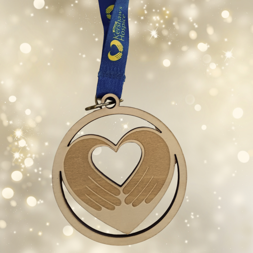 A circular wooden bauble with two hands in the shape of a heart, dangling by a dark blue ribbon with the Dr Kershaw's logo