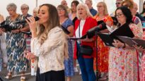A young woman with blonde hair singing in front of a choir dressed in colourful clothing