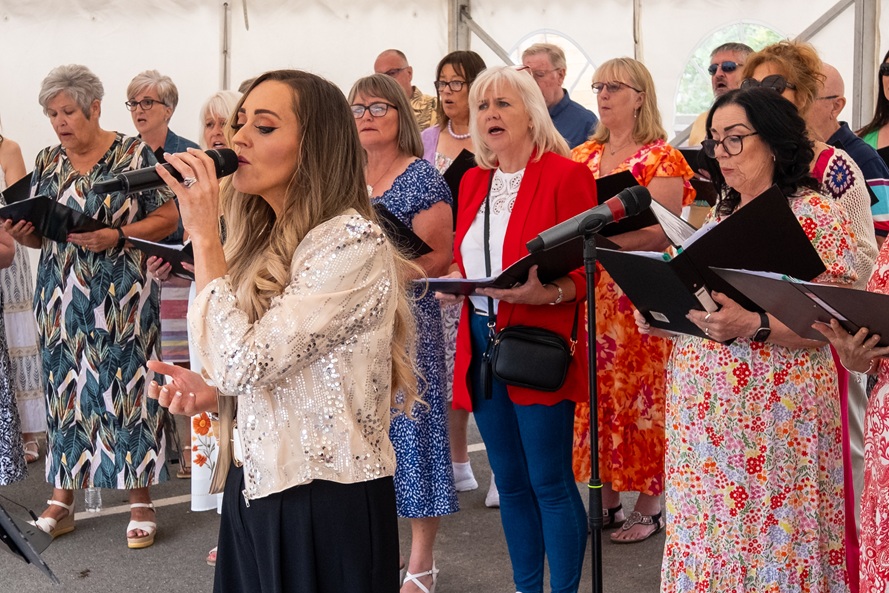 A young woman with blonde hair singing in front of a choir dressed in colourful clothing