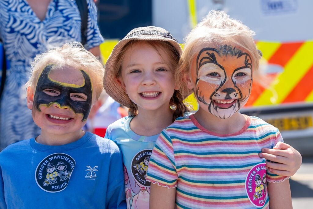 Three young children smiling - the one on the left has had her face painted as batman, and the one on the right has her face painted like a tiger