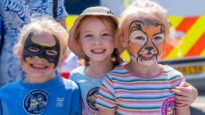 Three young children smiling - the one on the left has had her face painted as batman, and the one on the right has her face painted like a tiger