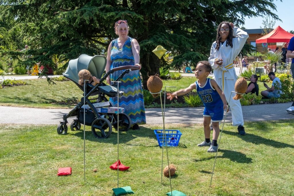 A family, including a woman with a pram, watching as a young boy plays the coconut shy in the garden