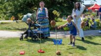 A family, including a woman with a pram, watching as a young boy plays the coconut shy in the garden