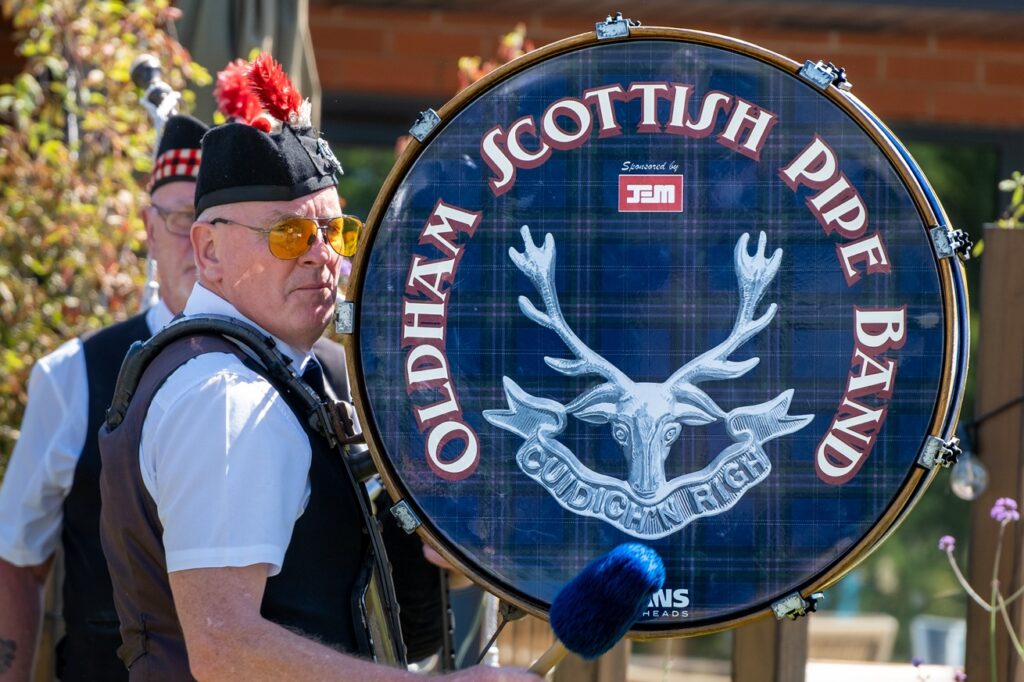 A man wearing a red feathered hat in front of a large drum saying 'Oldham Scottish Pipe Band'