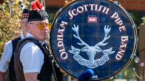 A man wearing a red feathered hat in front of a large drum saying 'Oldham Scottish Pipe Band'