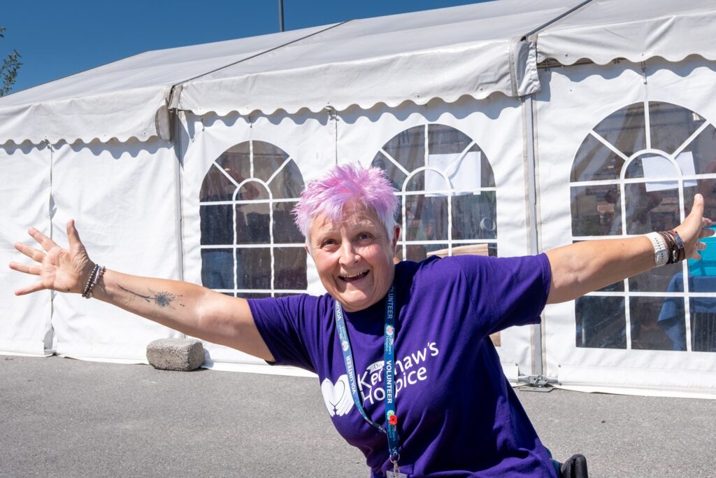 A woman in a wheelchair with short pink hair holding her arms out in front of a row of white marquees