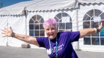 A woman in a wheelchair with short pink hair holding her arms out in front of a row of white marquees