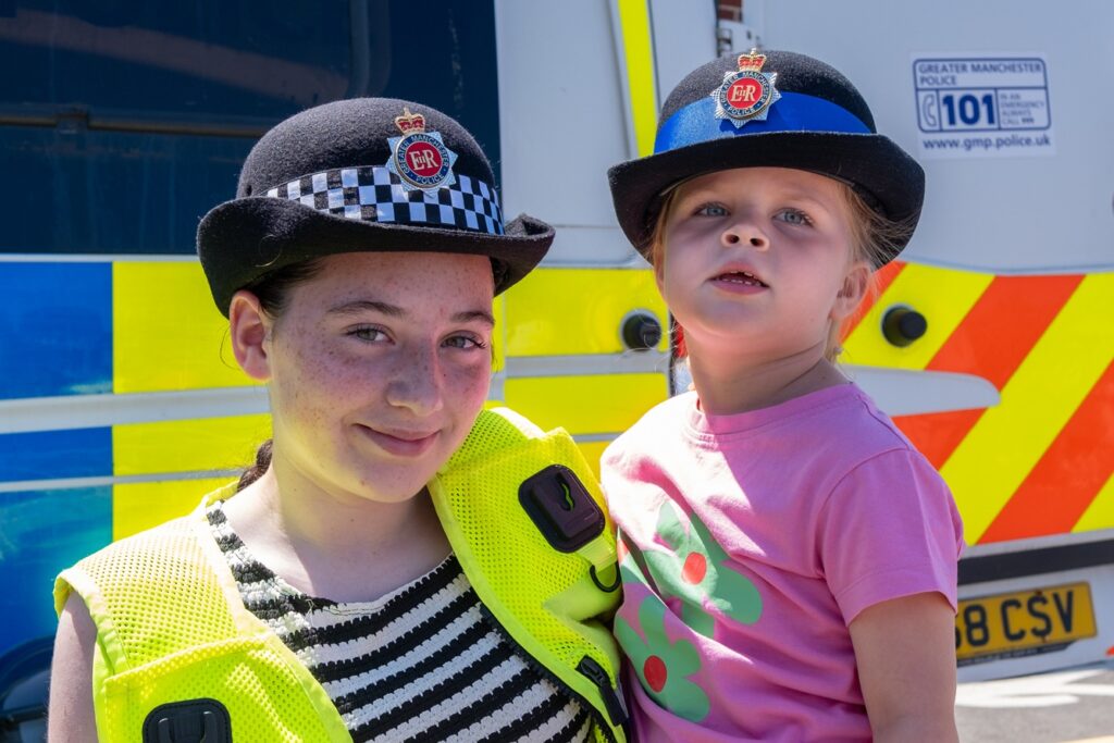 A young girl in a stripy t-shirt and high-vis jacket holding a toddler. They are both wearing police hats