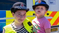 A young girl in a stripy t-shirt and high-vis jacket holding a toddler. They are both wearing police hats